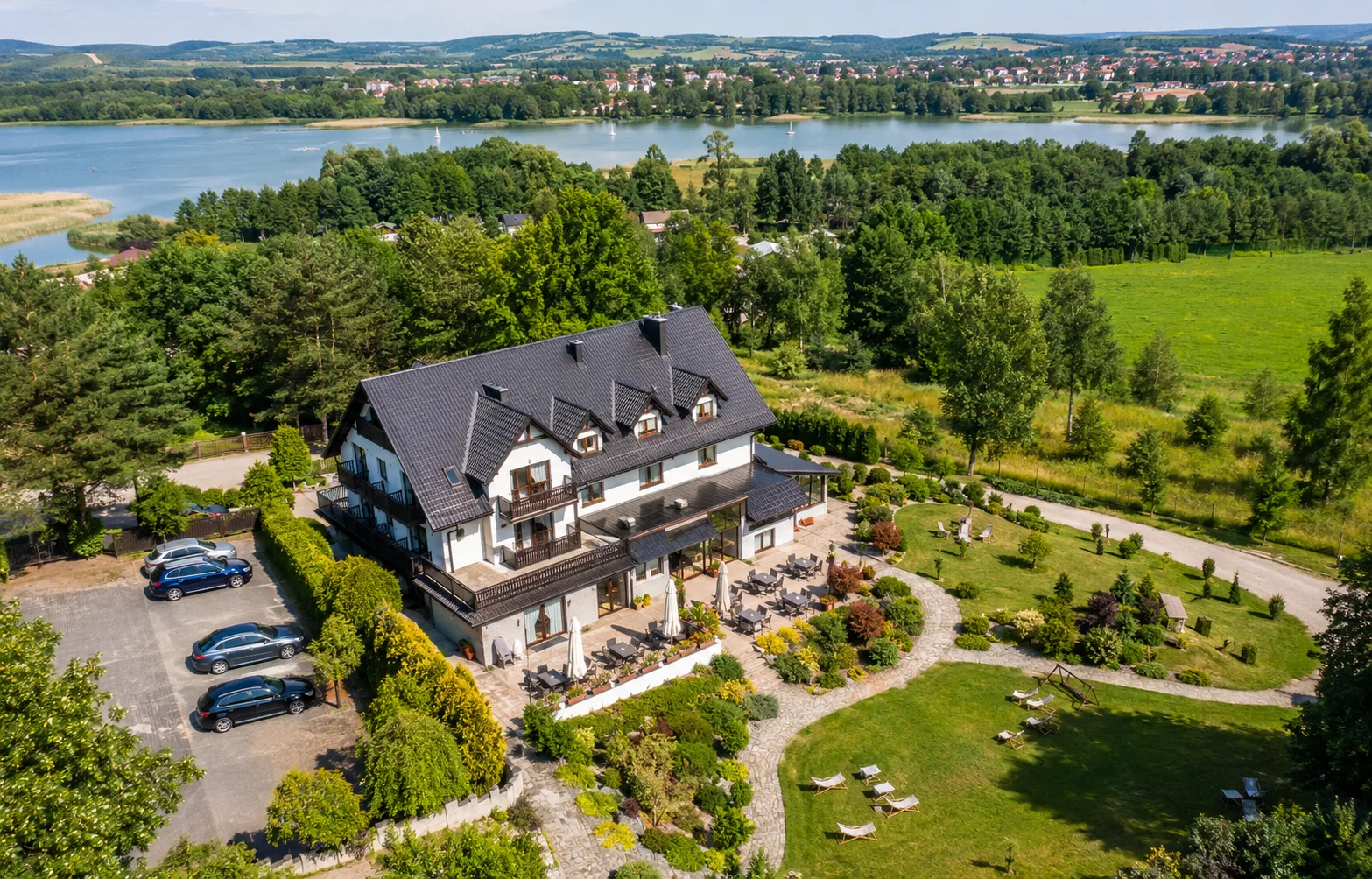 Hotel lub pensjonat pokazany z drona z widocznym ogrodem, parkingiem, jeziorem i okolicą